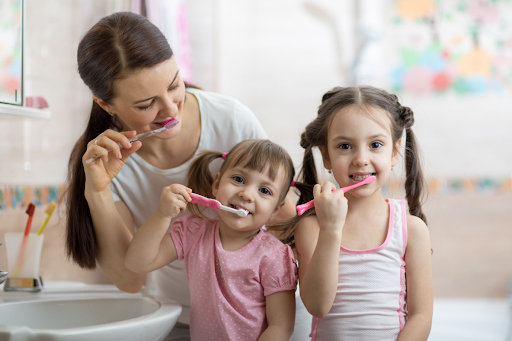 Two young girls in pink pajamas brushing their teeth in front of the bathroom sink with their mom